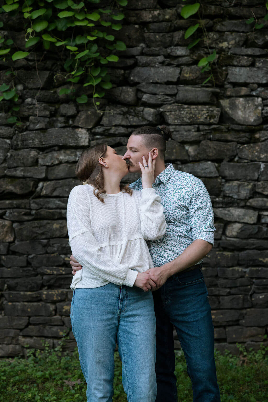 Couple sharing a kiss against a rustic stone wall covered in climbing greenery at Knoxville Botanical Garden, the woman in a cream sweater and jeans, the man in a floral button-down.