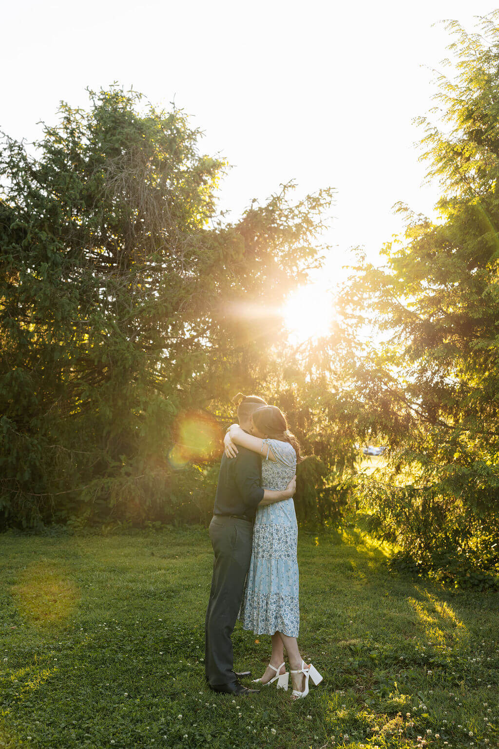 Couple embracing in an open lawn at Knoxville Botanical Garden during golden hour, the sun bursting through the tree line behind them and casting a warm glow across the scene.