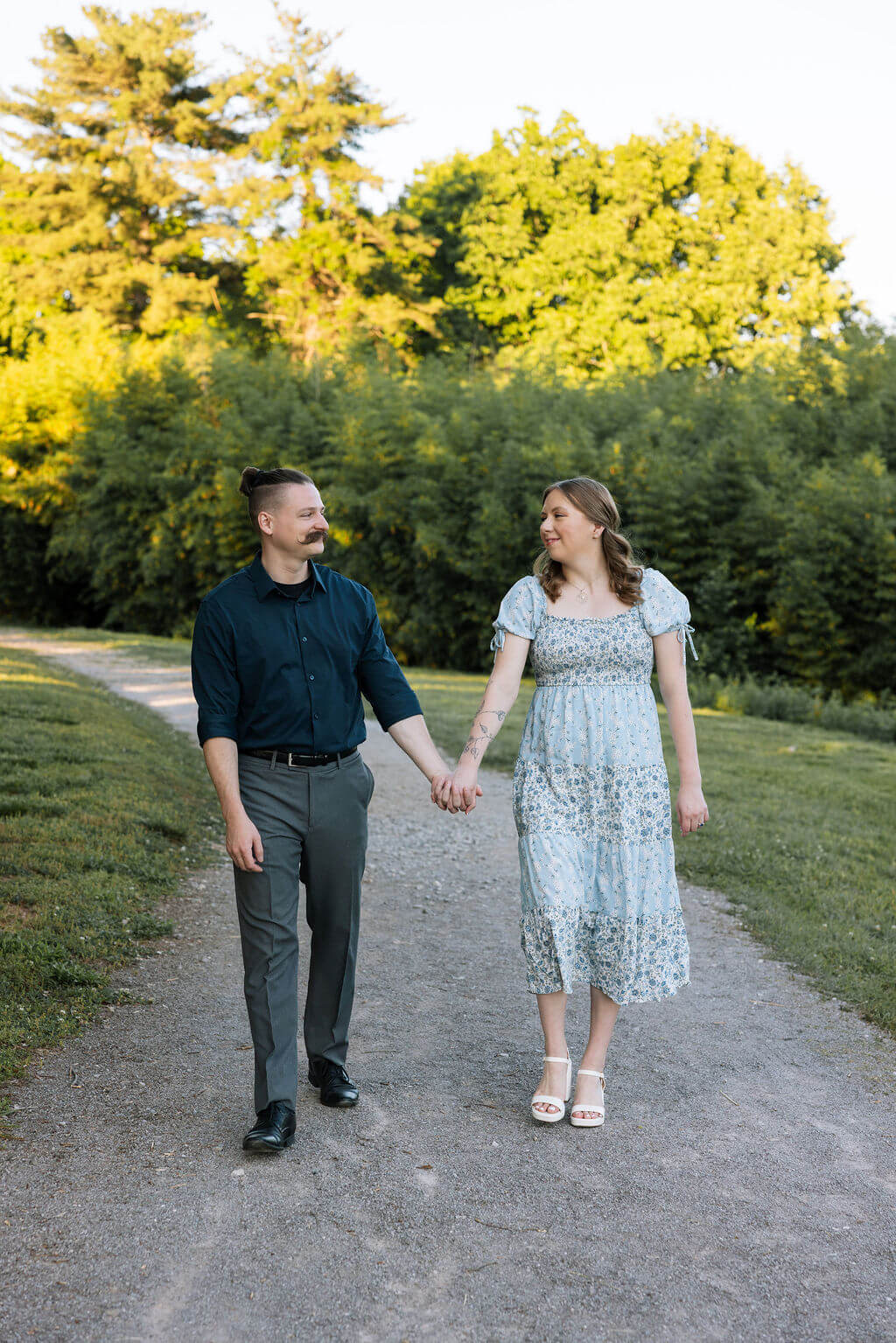 Engaged couple walking hand in hand down a gravel path at Knoxville Botanical Garden, laughing and looking at each other surrounded by lush green trees in summer.