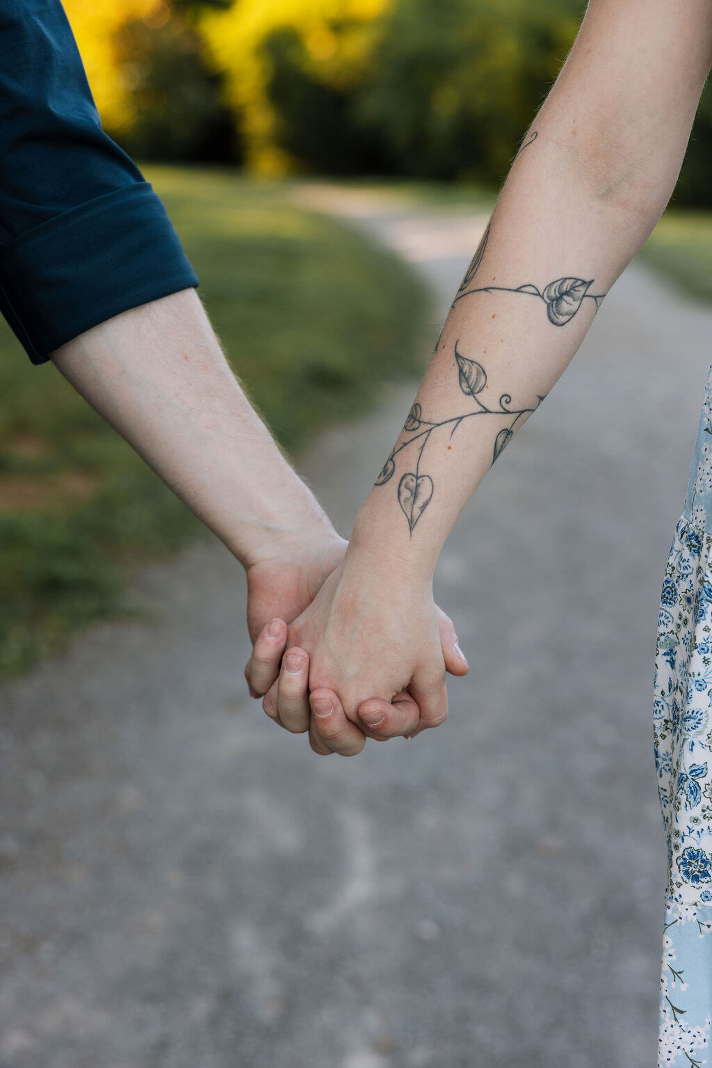 A close-up of a couple holding hands on a gravel path, the woman's wrist adorned with a delicate botanical vine tattoo, captured during an outdoor engagement session in Knoxville, Tennessee.