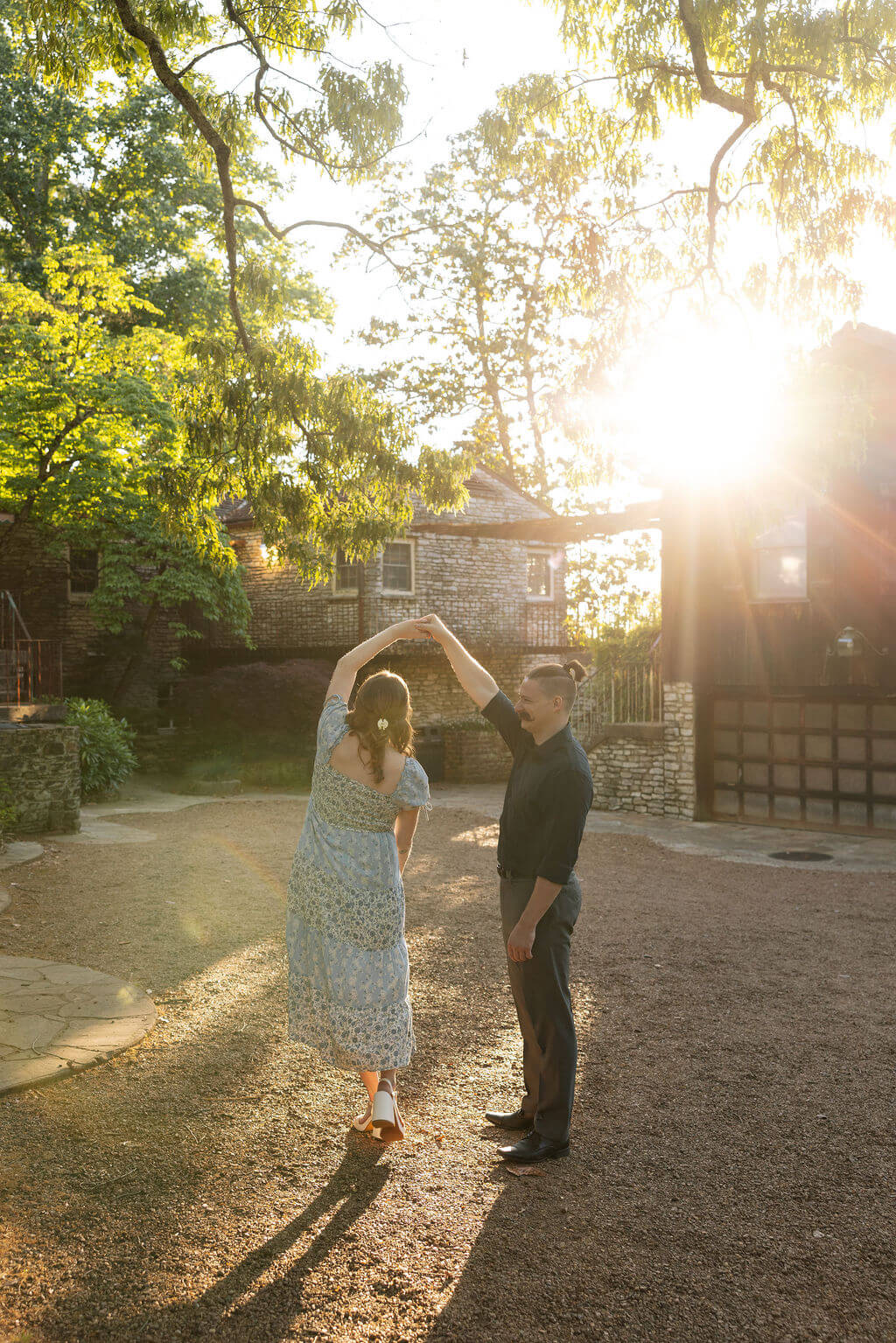 A couple dances together at golden hour, the woman in a flowy blue floral dress spinning as warm sunlight bursts through the trees behind them during a Knoxville engagement session.