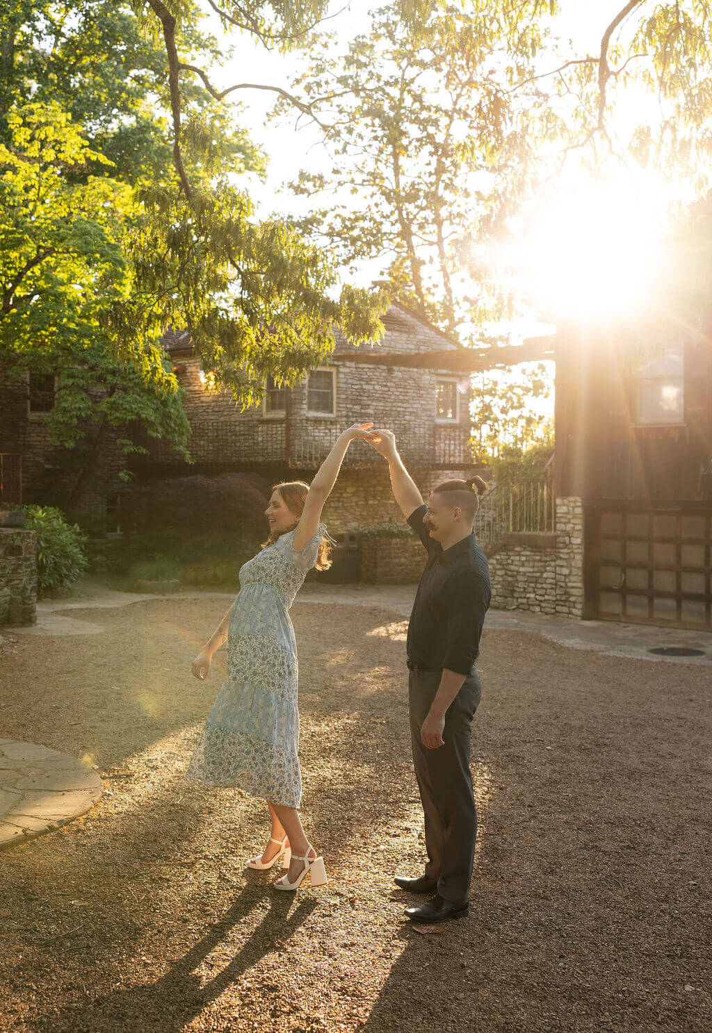 Couple dancing together on a gravel path at Knoxville Botanical Garden during sunset, the man spinning his partner under his arm as golden light floods the scene from behind the trees and stone buildings.