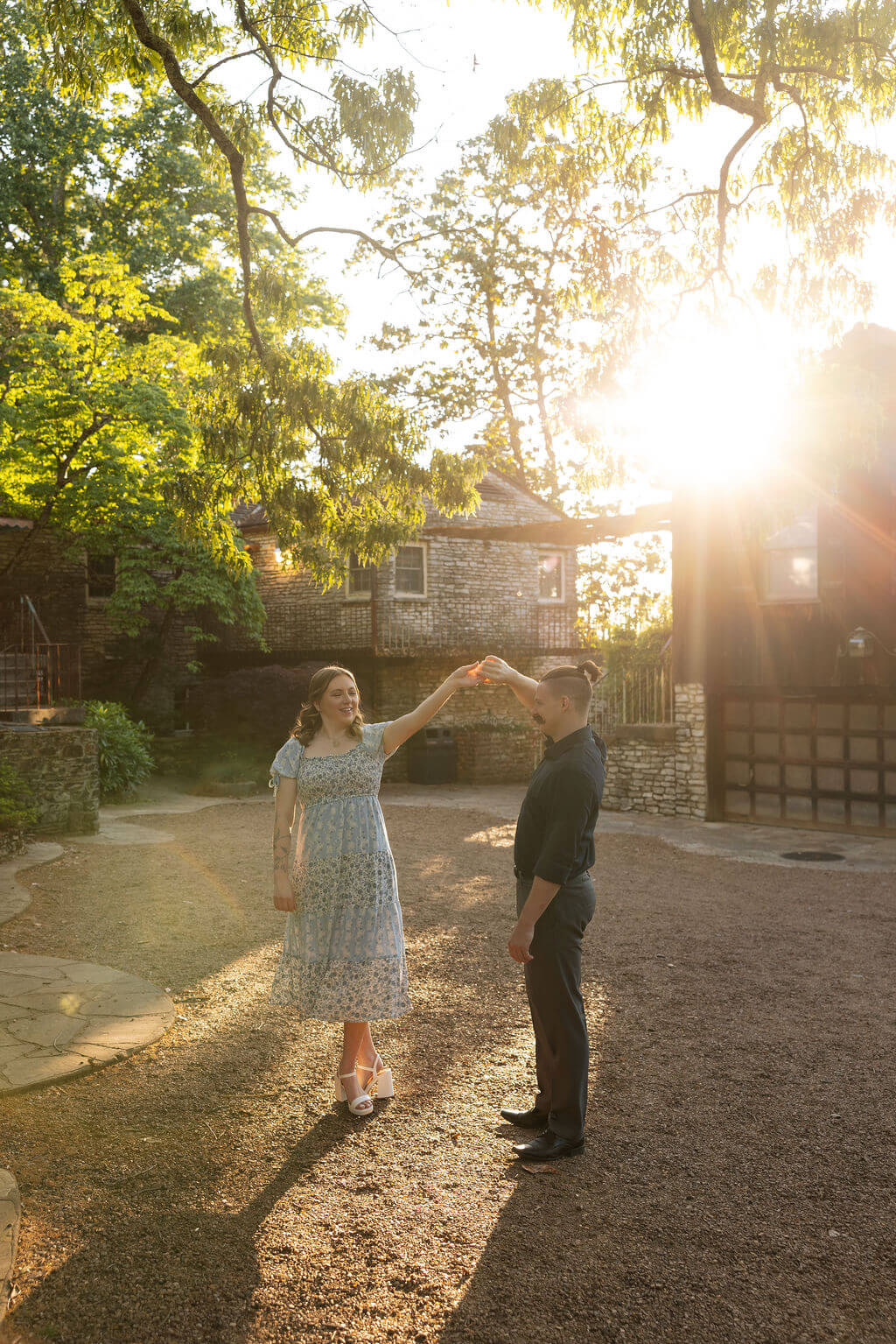 Engaged couple dancing on a gravel path at Knoxville Botanical Garden at golden hour, the woman spinning with her arm raised as a historic stone building and iron gate glow in the background.