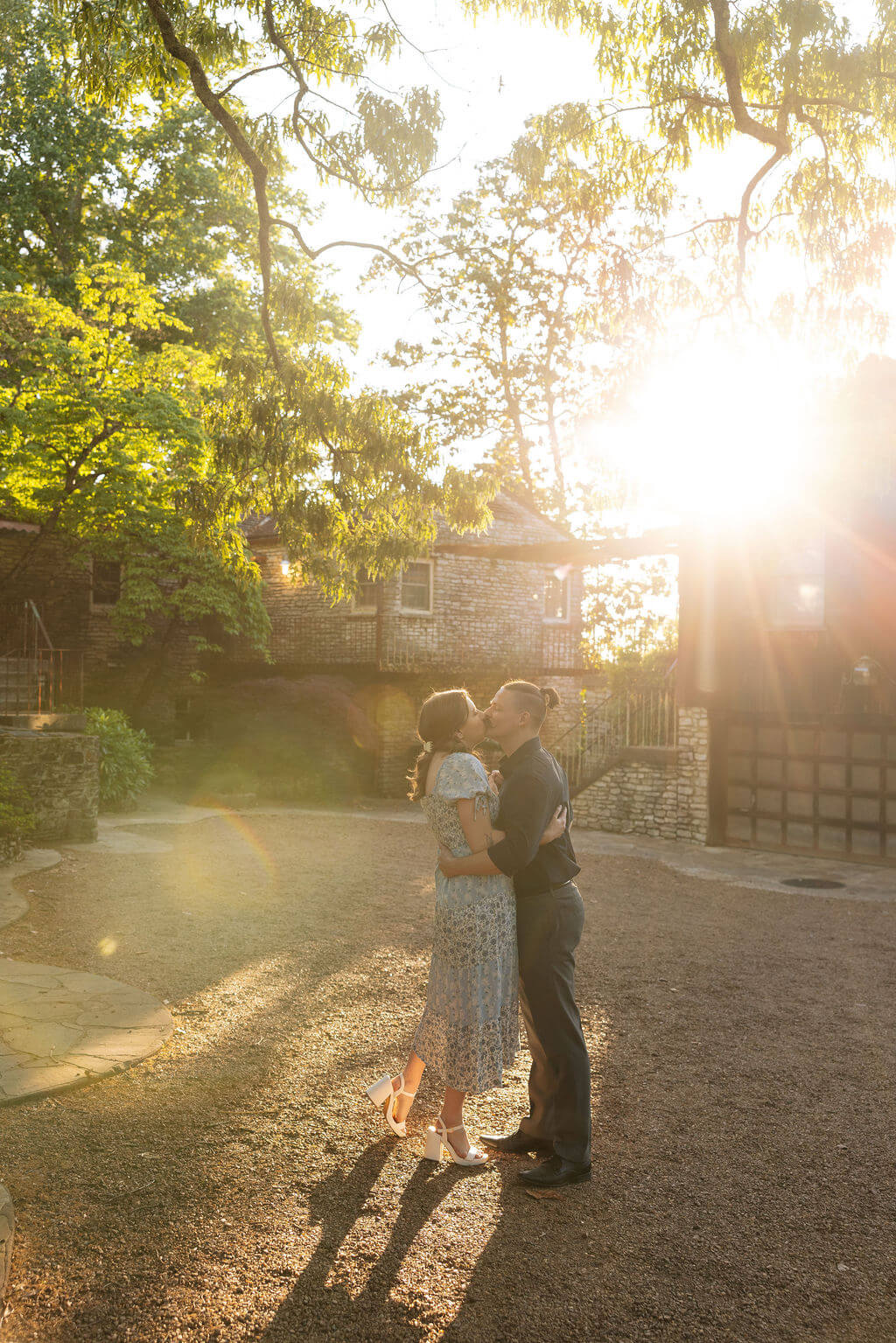 Couple pulling each other close on a gravel path at Knoxville Botanical Garden at sunset, about to kiss, with dramatic golden sun flare and a historic stone building glowing warmly behind them.