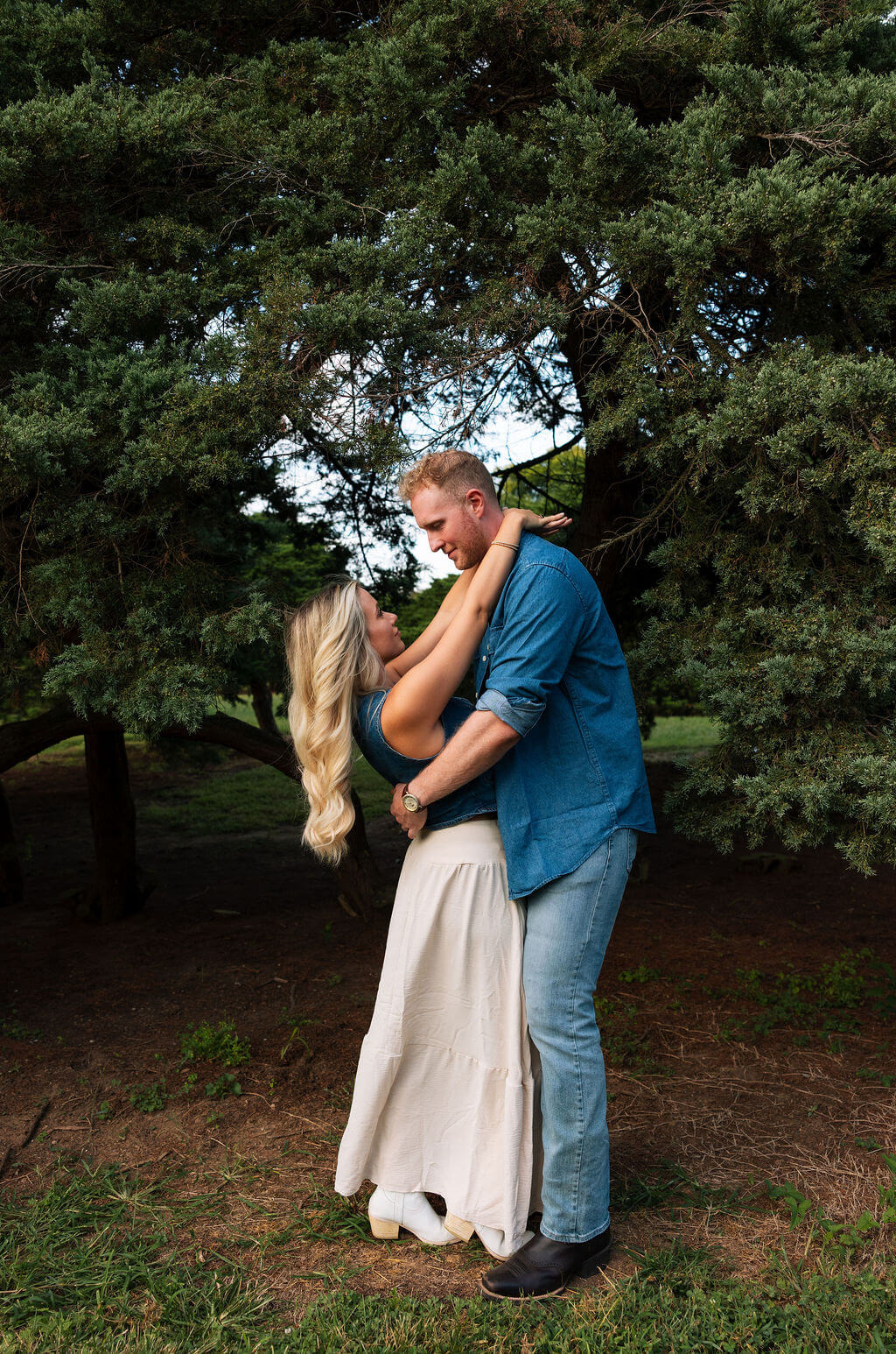 Couple embracing beneath a large evergreen tree during an outdoor engagement session. The woman wears a cream skirt and white cowboy boots while looking up at her partner in a denim shirt and jeans.