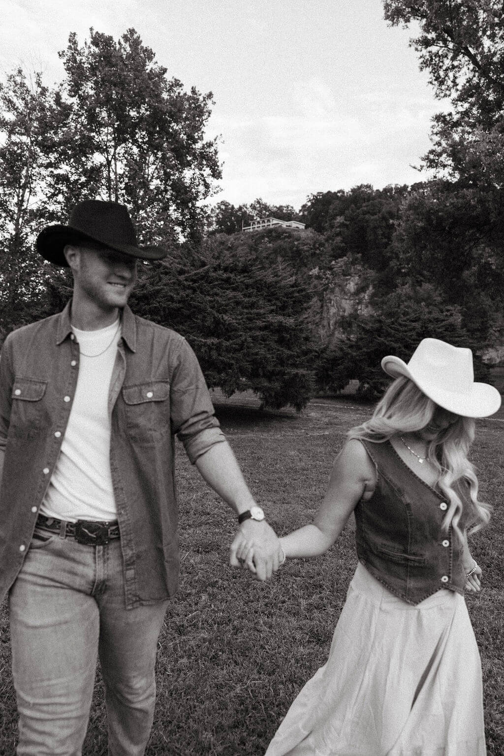 Black and white engagement photo of a couple walking hand in hand across a grassy field while smiling at one another.