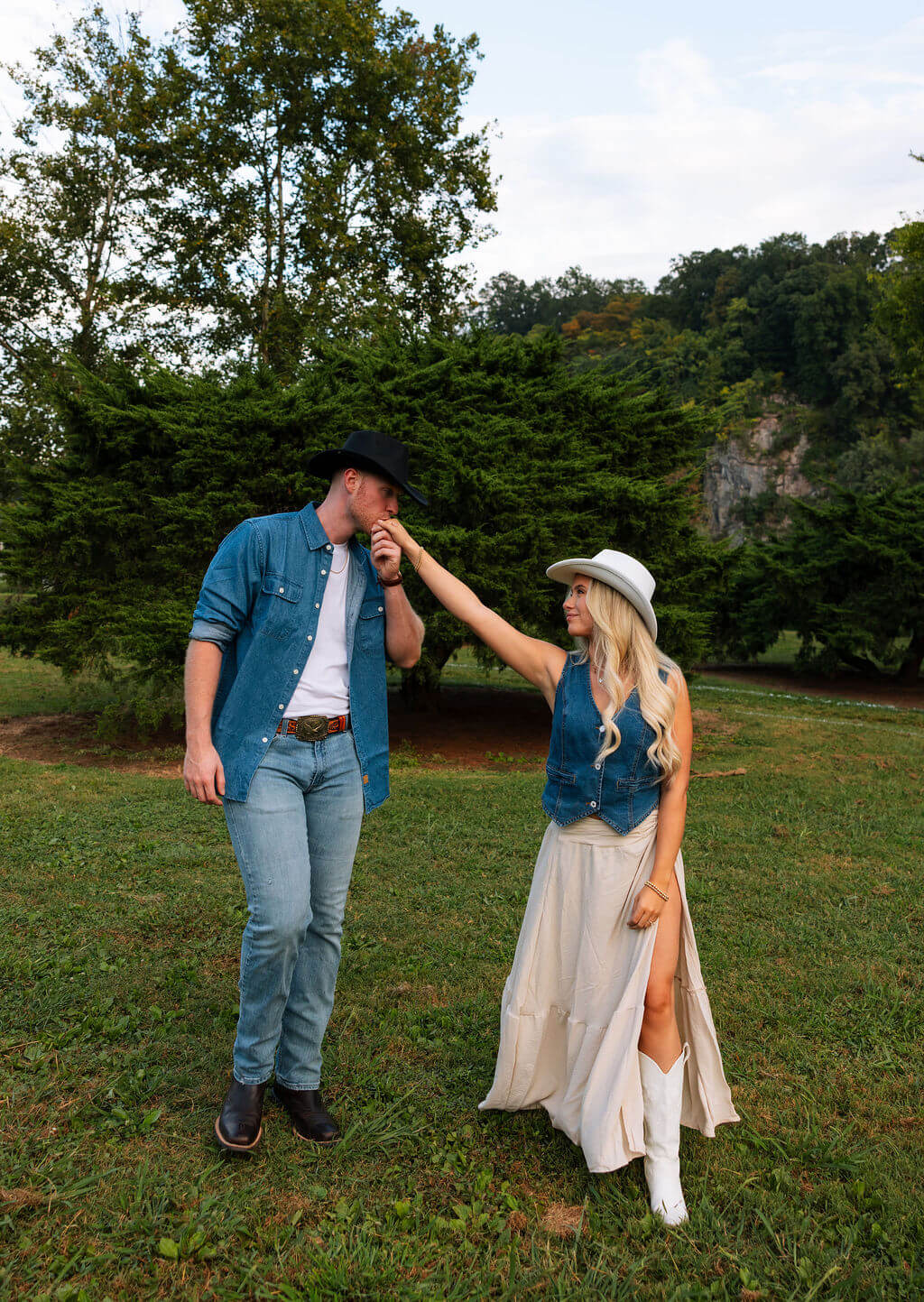 Groom-to-be kissing his partner’s hand during an outdoor engagement session. The couple stands in a grassy field wearing coordinating western-inspired outfits.