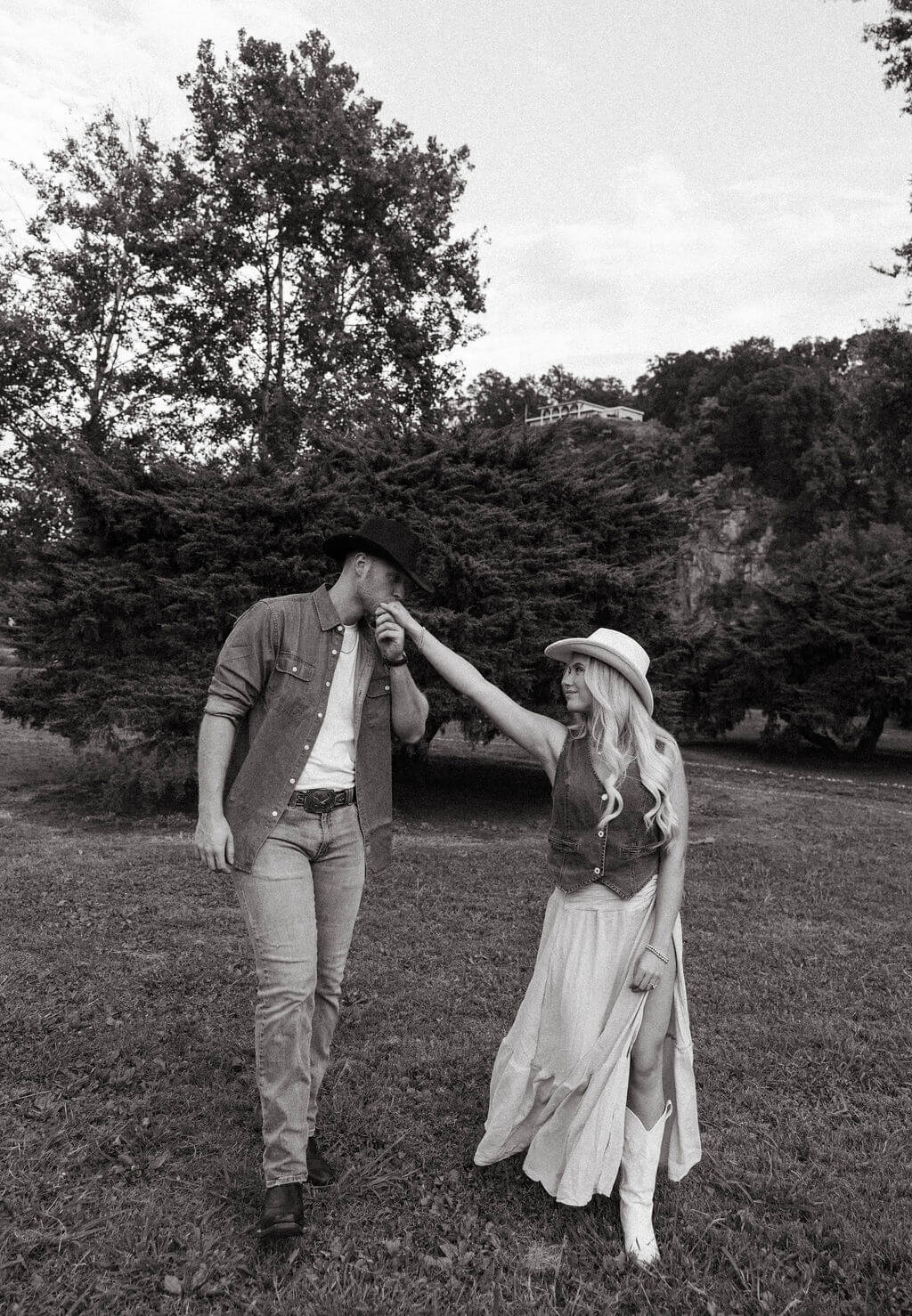 Black and white photo of a groom kissing his partner’s hand while they stand together in a grassy field.
