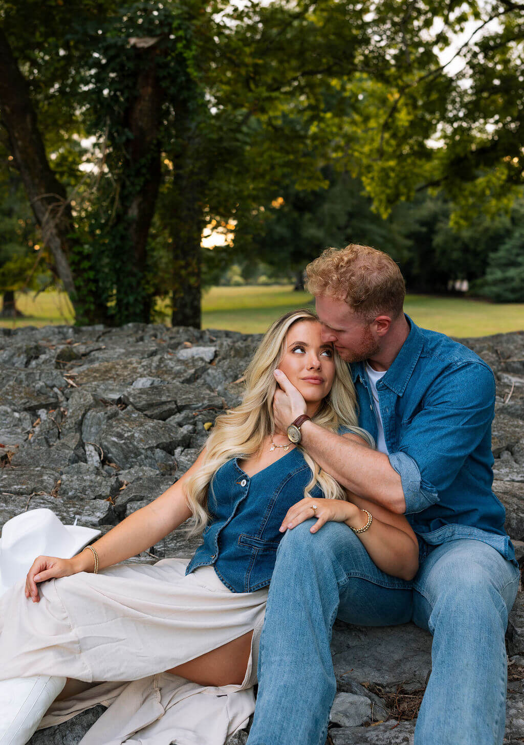 Couple sitting together on a stone wall during an engagement session. The groom rests his head against his partner while she looks toward the camera.