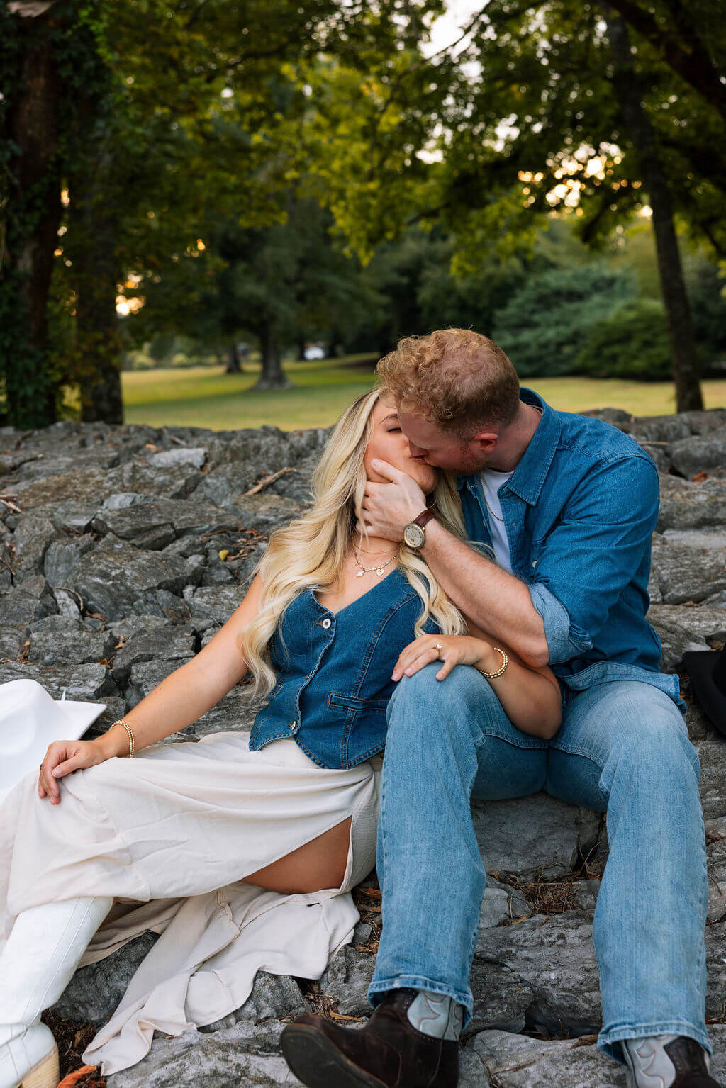 Couple sitting together on a rocky stone wall at sunset. The groom leans in to kiss the bride-to-be while she sits in a cream skirt and denim vest.