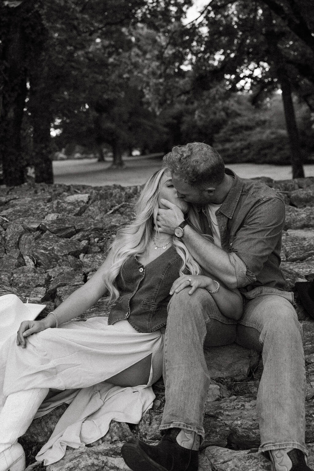 Black and white engagement portrait of a couple kissing while seated on a rocky stone wall.