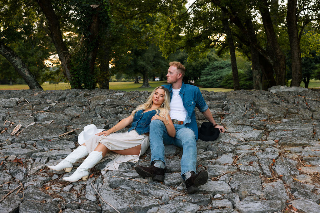 Couple relaxing on a stone wall in a park surrounded by mature trees. The woman reclines against her partner while both look off into the distance.