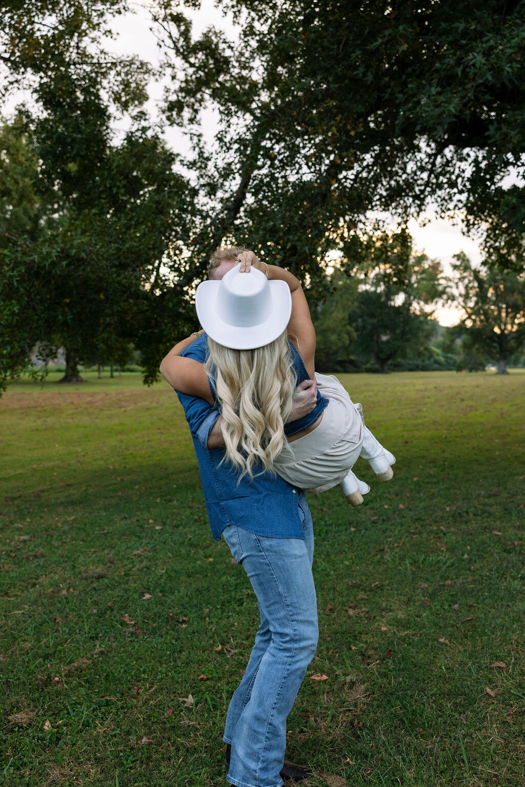 Groom carrying his partner across a grassy field while she wears a white cowboy hat and cream skirt.