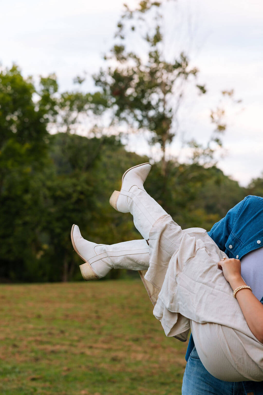 Detail photo of a bride-to-be’s white cowboy boots and flowing cream skirt as her partner carries her during an engagement session.