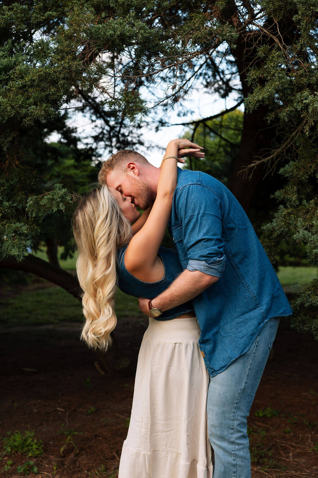 Close-up engagement portrait of a couple embracing beneath a tree. The bride-to-be wraps her arms around her partner’s neck as they lean in forehead to forehead.