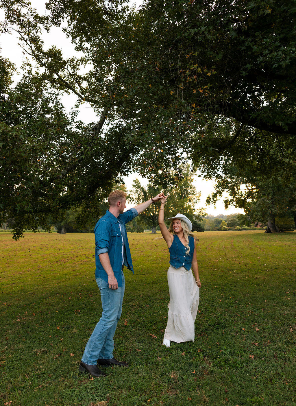 Groom twirling his partner beneath a large tree during an outdoor engagement session in a grassy park.