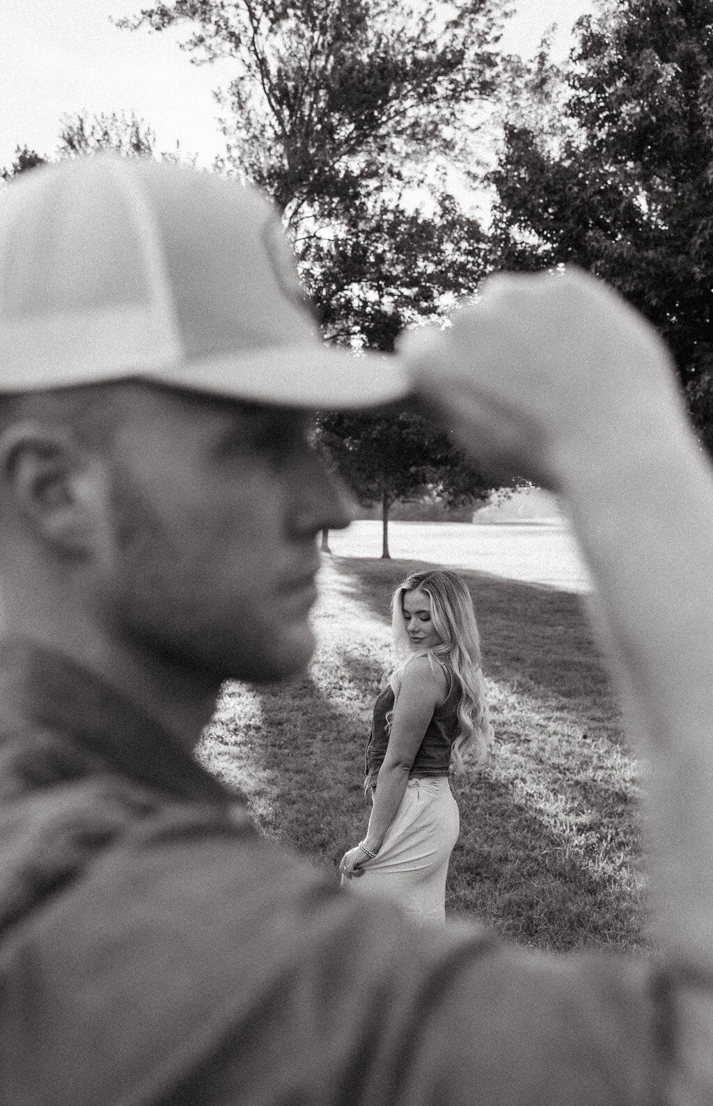 Artistic black and white portrait with the groom in the foreground adjusting his hat while the bride-to-be stands softly blurred in the background.