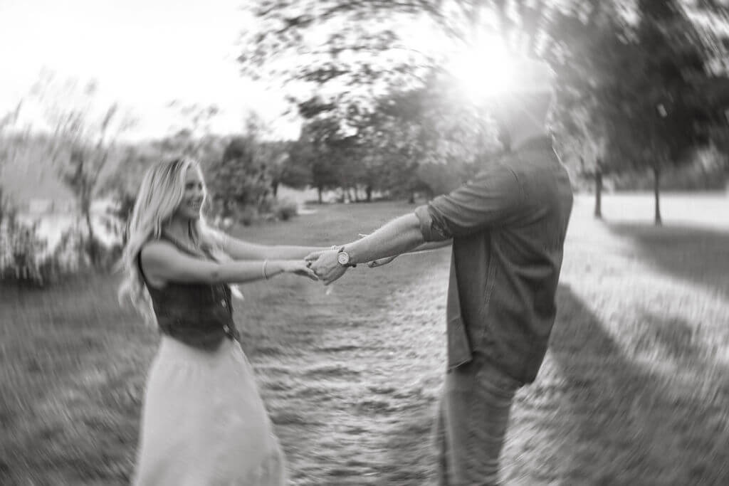 Artistic black and white photo of a couple spinning together while holding hands. Motion blur surrounds them as sunlight filters through the trees.