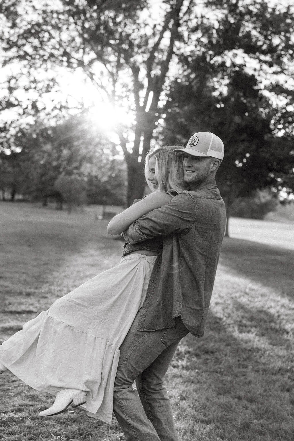 Black and white photo of a groom lifting his partner during an outdoor engagement session. Sunlight filters through the trees as the couple smiles together.