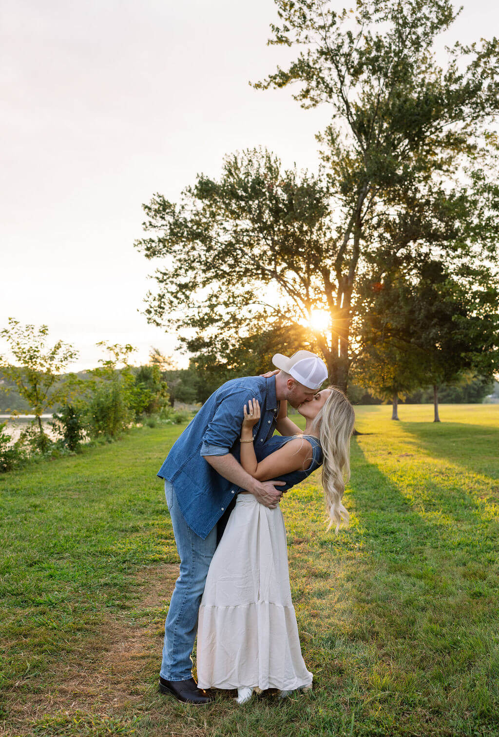 Couple sharing a kiss during golden hour beside a tree-lined path. The setting sun creates warm backlighting behind them.