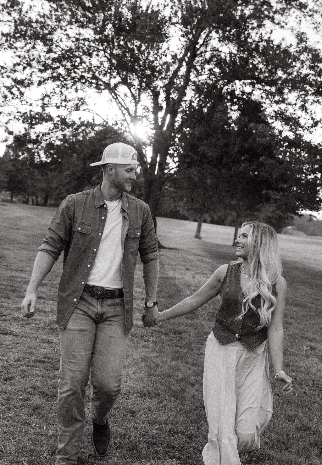 Black and white engagement photo of a couple walking hand in hand through a grassy park beneath large trees.