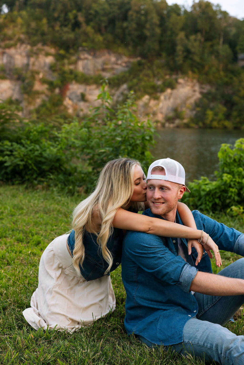 Couple sitting together on the grass near a lakeside shoreline during an outdoor engagement session.