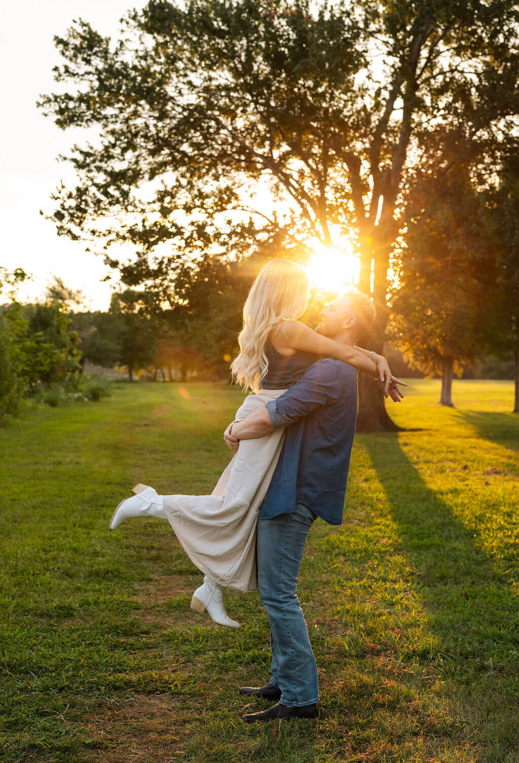 Couple embracing during golden hour with sunlight streaming through the trees behind them. The groom lifts his partner off the ground as they hug.