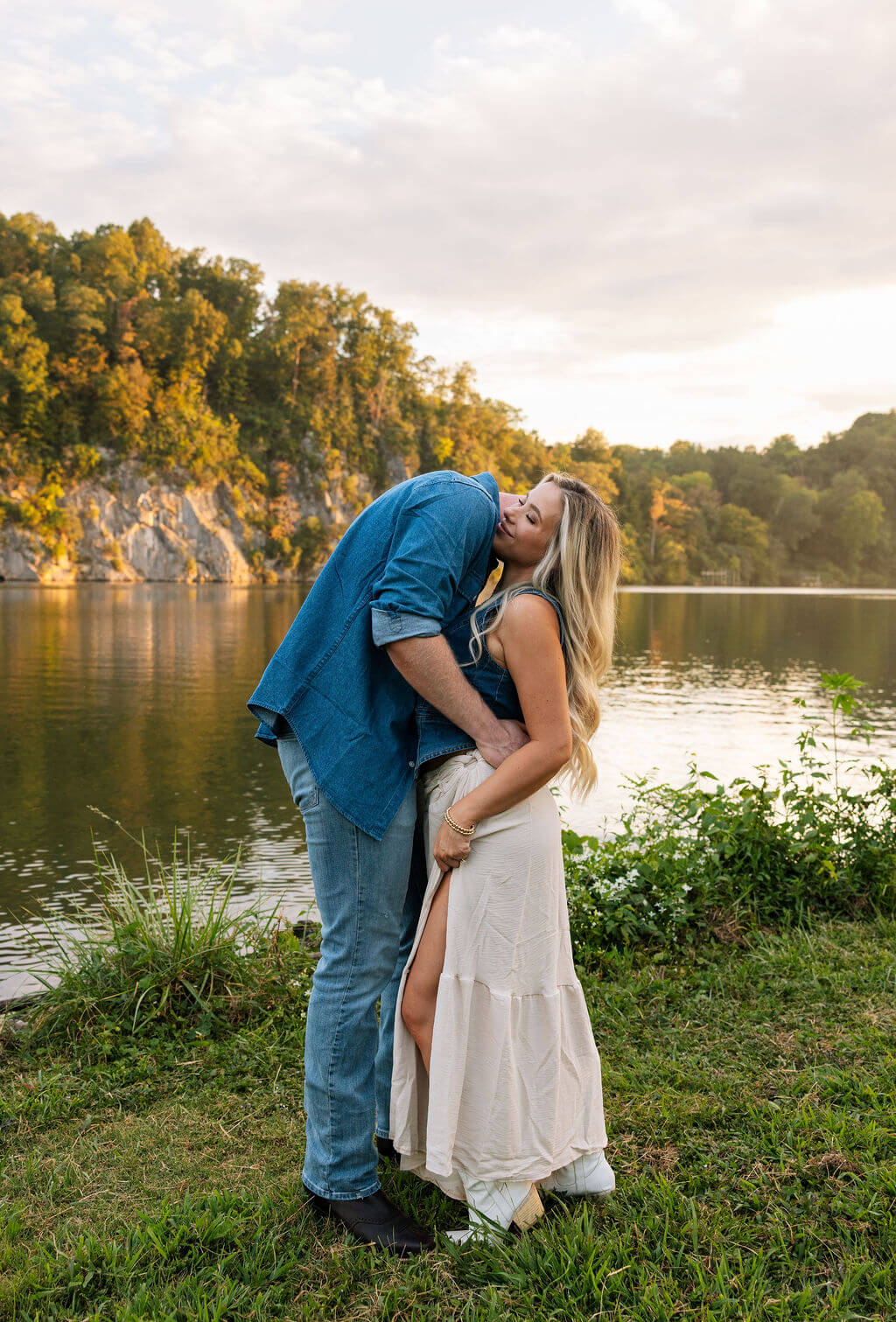 Couple kissing beside a lake during an outdoor engagement session. Rocky cliffs and calm water create a scenic backdrop.