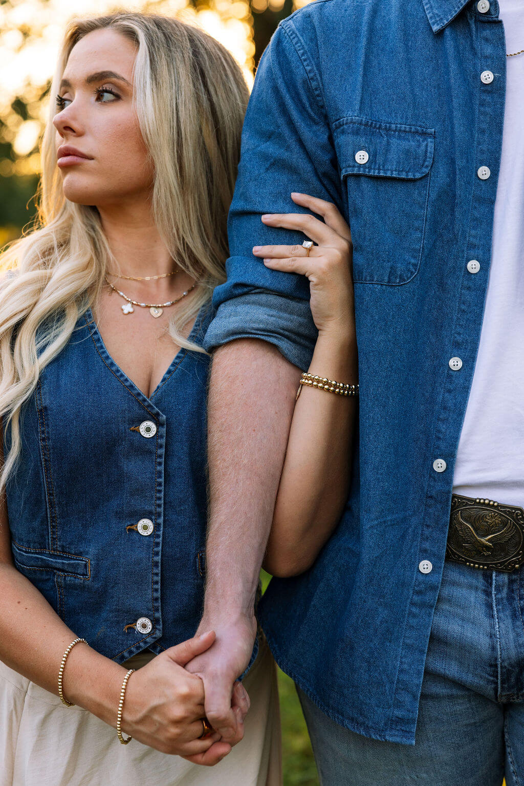 Close-up engagement photo of a couple holding hands. The bride-to-be rests her hand on her partner’s arm while wearing layered gold jewelry.