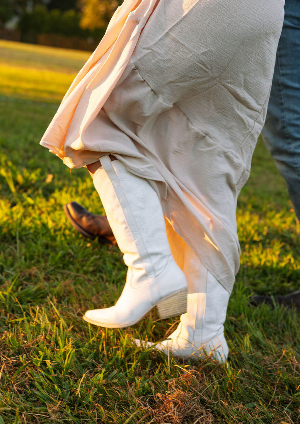 Detail photo of white cowboy boots peeking out beneath a flowing cream skirt during a sunset engagement session.