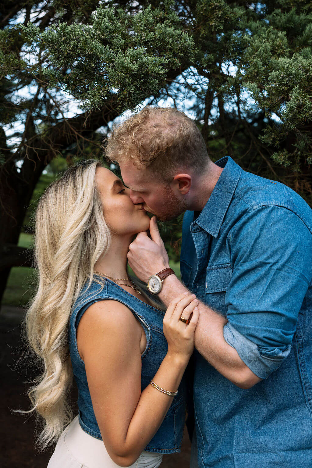 Close-up portrait of a couple sharing a kiss beneath an evergreen tree during an outdoor engagement session.