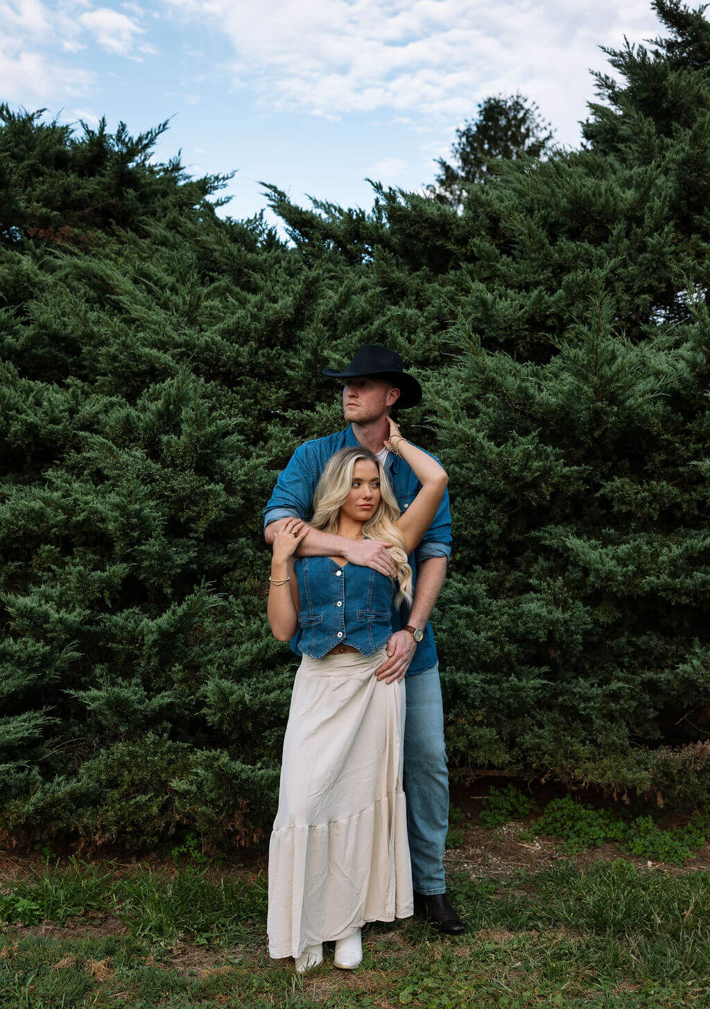 Couple standing together in front of tall evergreen trees during an outdoor engagement session. Both wear denim and western-inspired accessories.