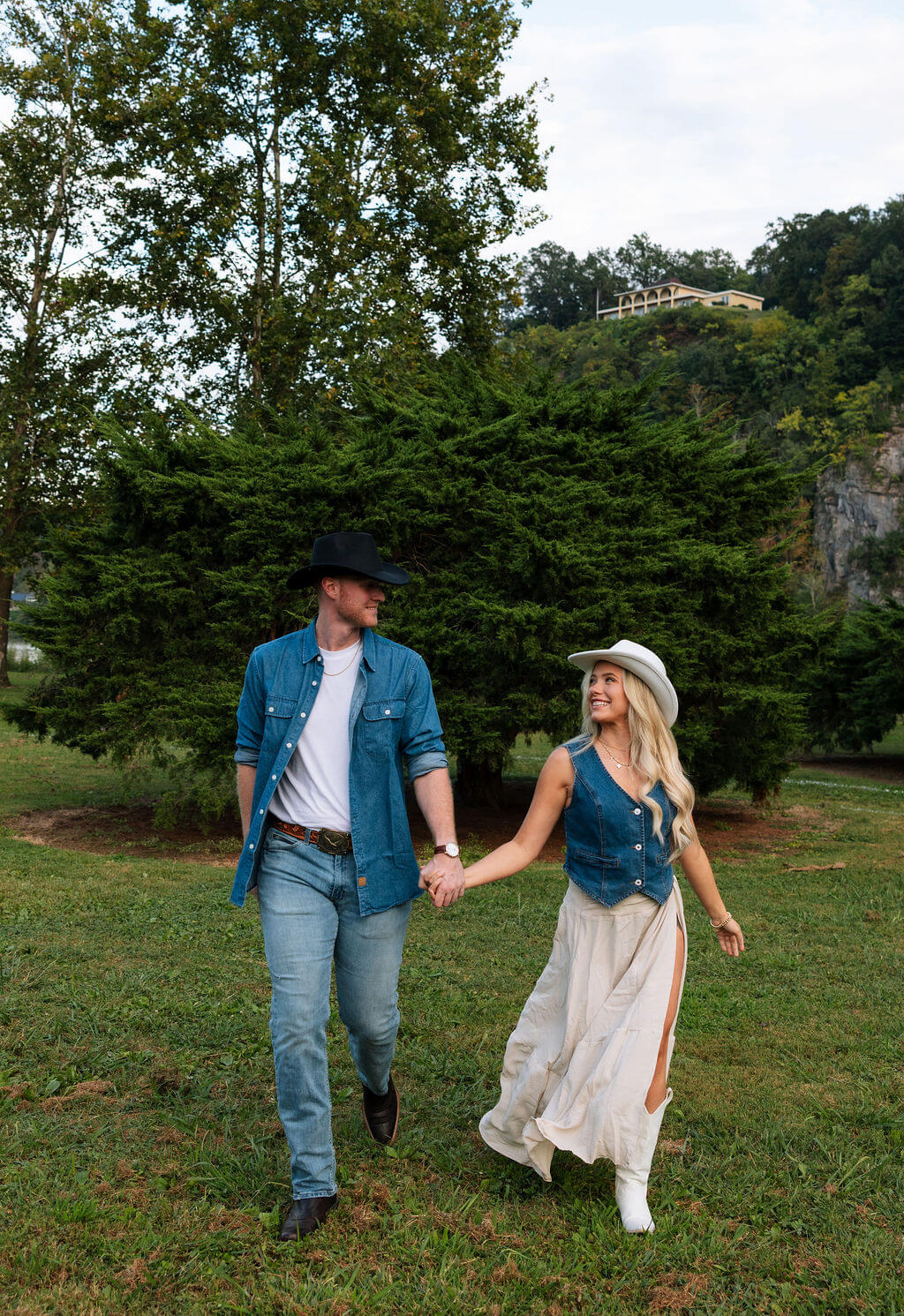 Engaged couple holding hands and walking across a grassy field during a countryside engagement session. Both wear western-inspired outfits with cowboy hats and denim.
