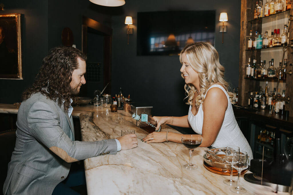 Woman in a white dress pours whiskey for her fiancé across a marble counter during a relaxed engagement photoshoot at one of the moody Knoxville bars.