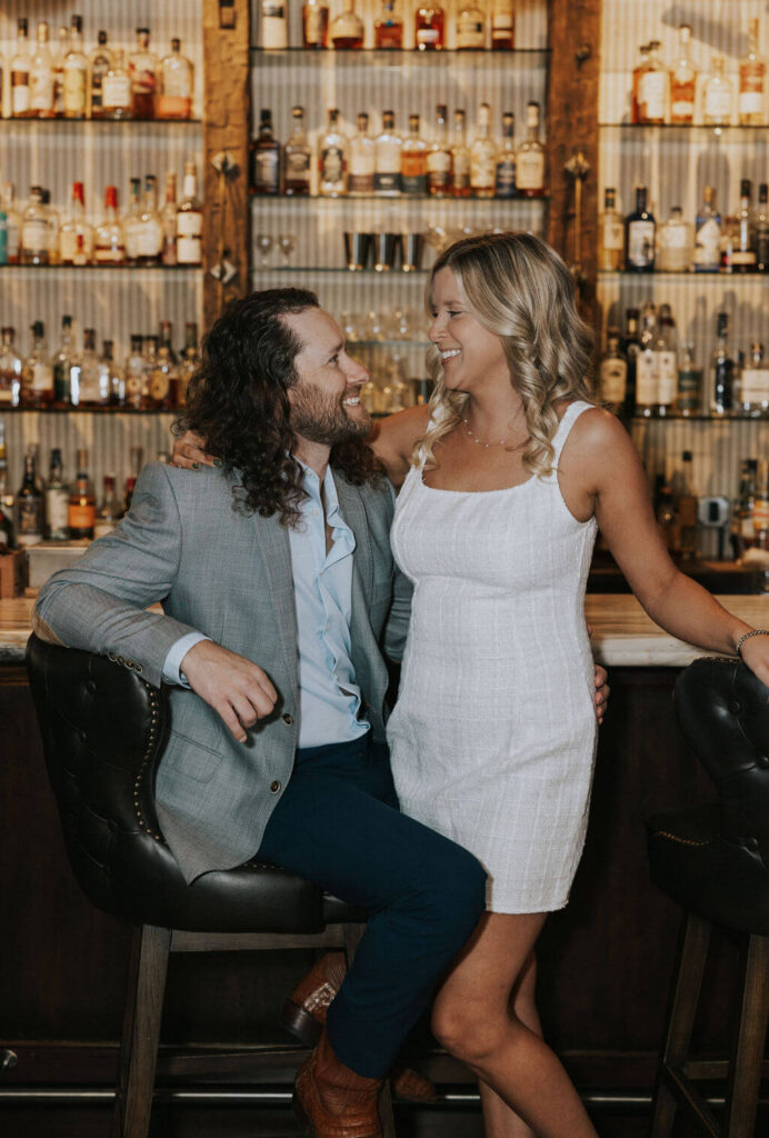 Couple laughing together beside shelves of liquor bottles during their relaxed engagement photos inside a Knoxville bar.