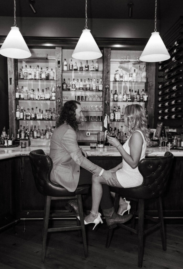 Black-and-white engagement photo of a couple sitting on leather barstools, holding hands and talking in front of shelves filled with liquor bottles.