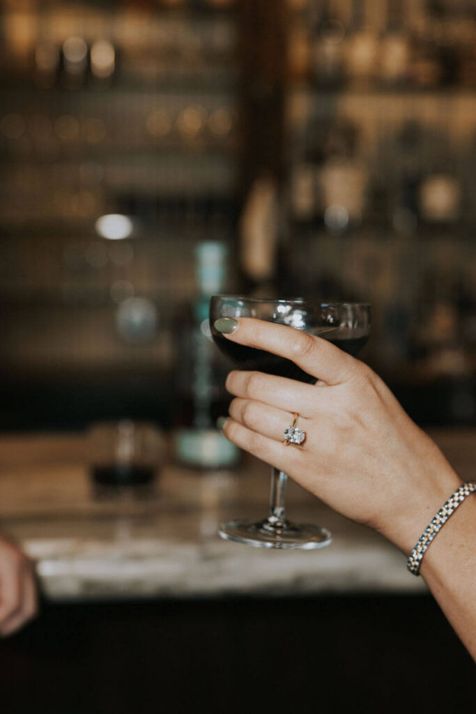 Close-up engagement photo of a woman’s hand holding a cocktail, with her engagement ring visible against the softly blurred bar background.