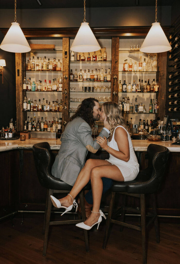 Couple sharing a kiss while seated together at a warmly lit Knoxville bar during their stylish indoor engagement photoshoot.