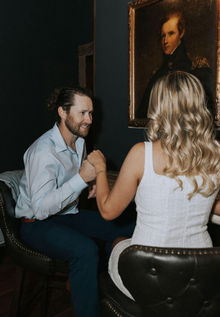Man gently kisses his fiancée’s hand as they sit across from each other on leather barstools during their engagement photoshoot.