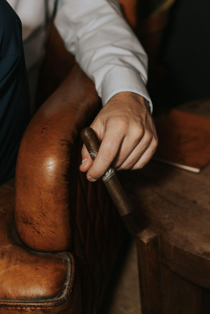 Close-up engagement photo of a man holding a cigar while resting his hand on the arm of a worn brown leather chair.