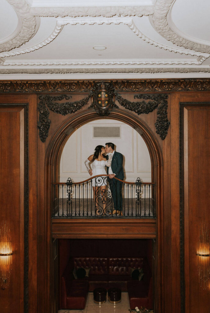 Engaged couple kissing on an interior balcony framed by an ornate wooden arch during their romantic engagement photos.