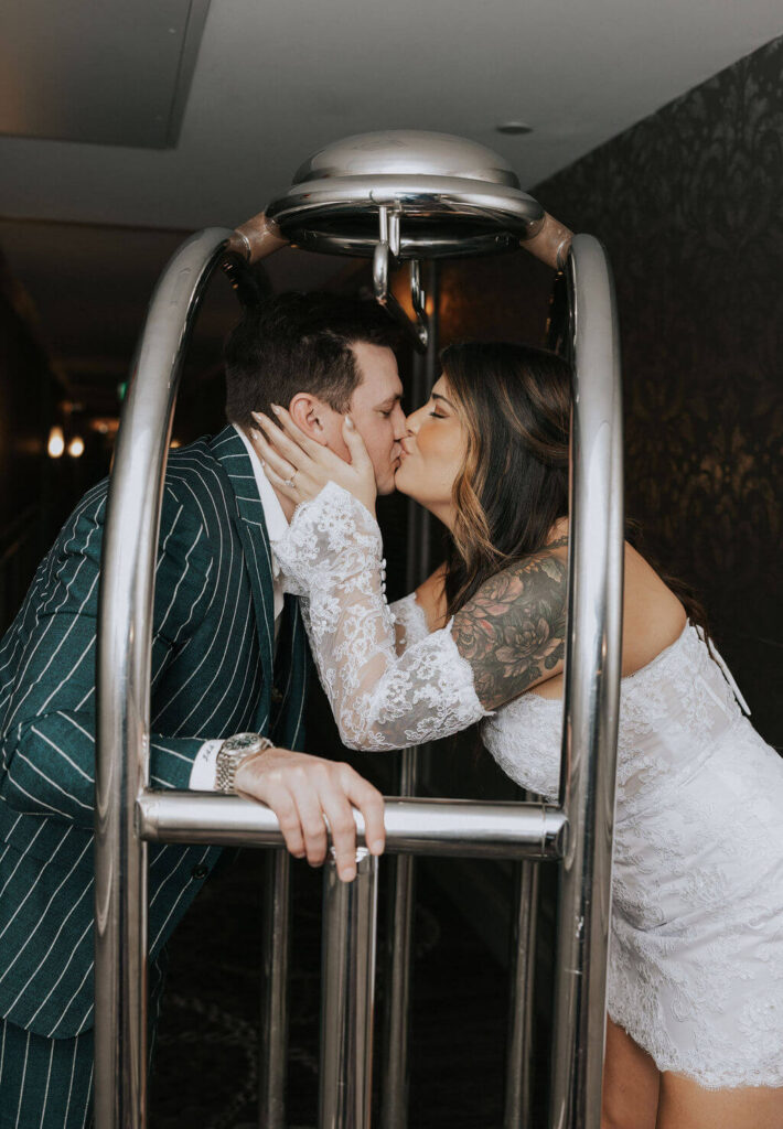 Couple in a green pinstripe suit and white lace dress kissing through a silver luggage cart during their playful engagement photoshoot.