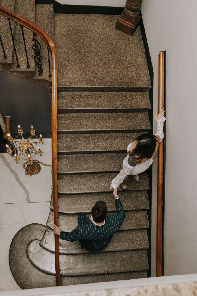 Overhead view of an engaged couple reaching for each other’s hands on a curved staircase inside a historic Knoxville hotel.