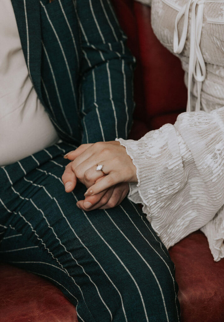 Close-up of an engaged couple holding hands on a red leather seat, with the woman’s diamond ring visible against the man’s green pinstripe suit.