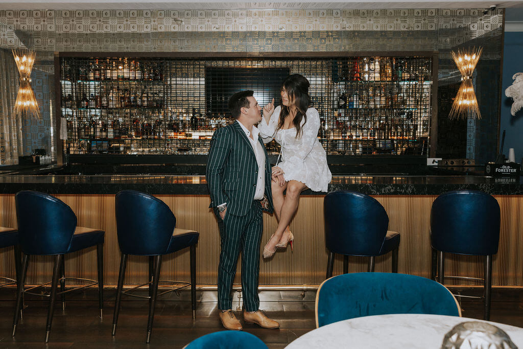 Woman in a white lace dress sitting on a bar counter while her fiancé stands beside her during an engagement photoshoot at one of the elegant Knoxville bars.
