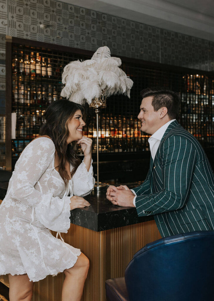 Couple smiling at each other across a dark bar counter during an elegant engagement photoshoot at one of the stylish Knoxville bars.