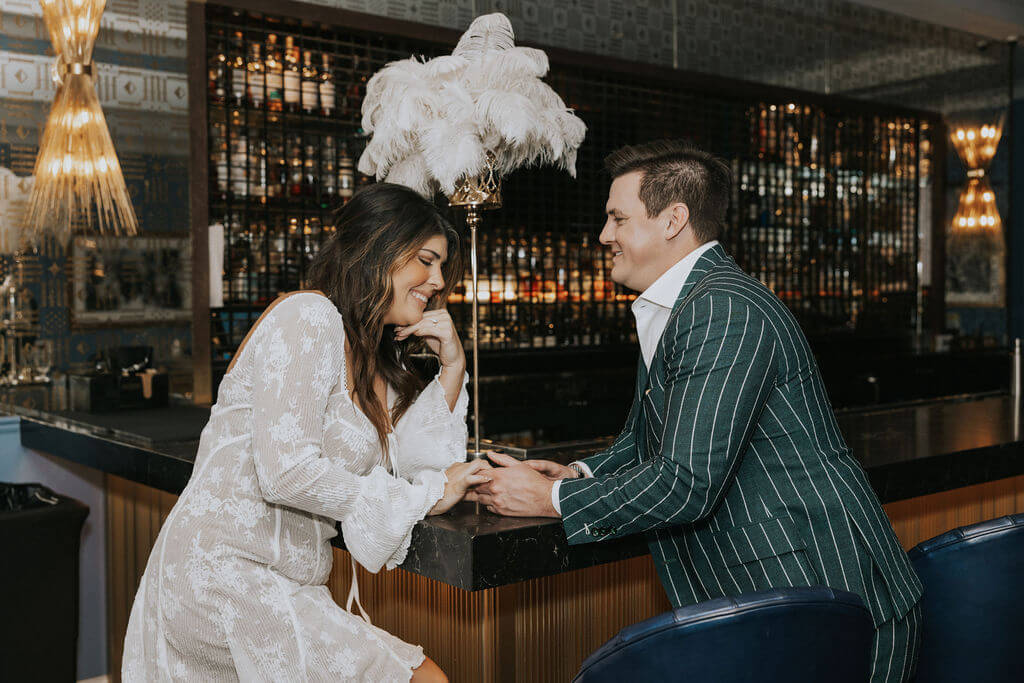 Engaged couple holding hands and laughing across a polished bar counter, with the woman in a lace dress and the man in a green pinstripe suit.