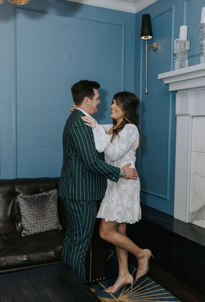 Engaged couple embracing beside a black leather sofa and blue paneled wall during their indoor Knoxville engagement photoshoot.