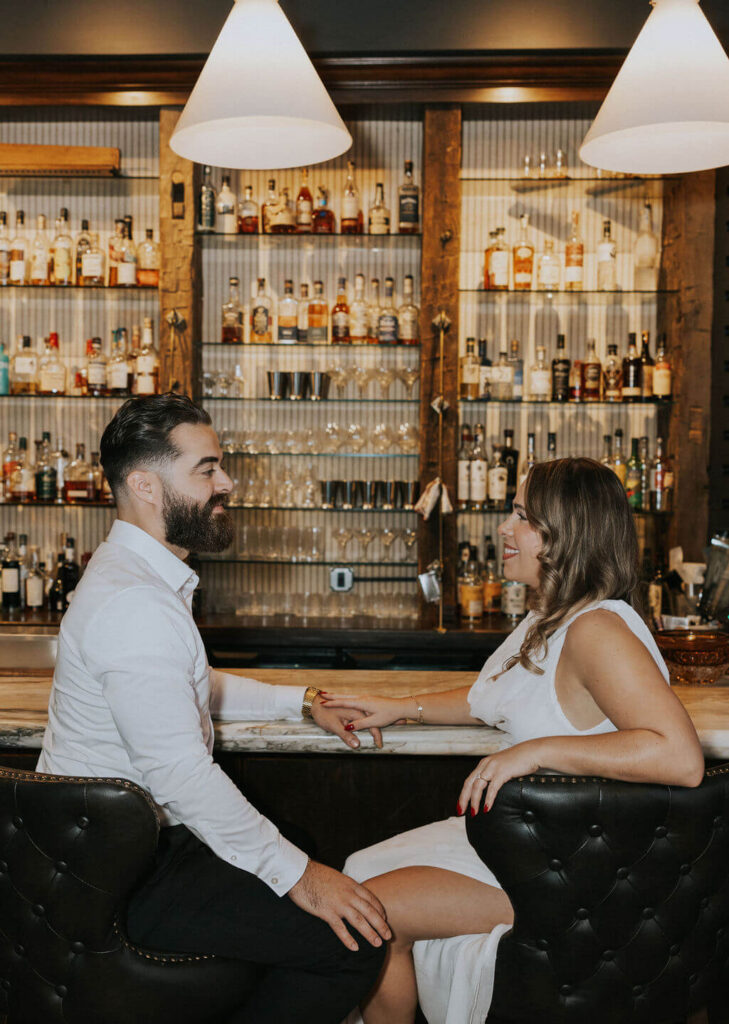 Engaged couple in white outfits sitting across from each other on black leather barstools and holding hands during an engagement photoshoot inside a warmly lit Knoxville bar.
