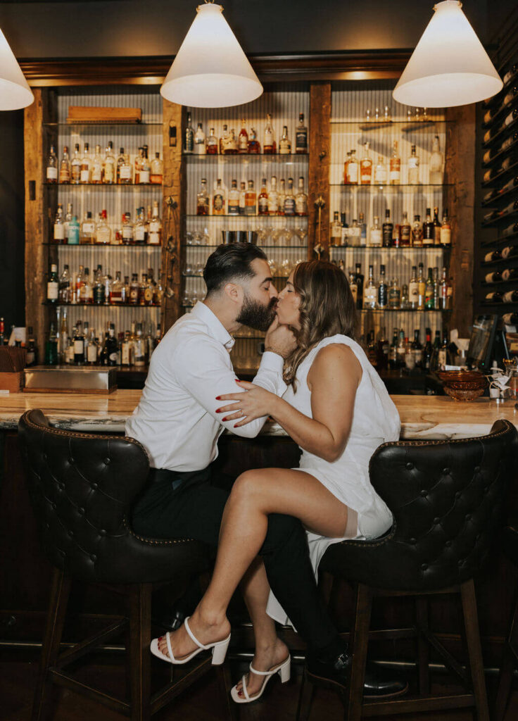 Engaged couple kissing on leather barstools in front of illuminated liquor shelves, with the woman wearing a white mini dress and heels.